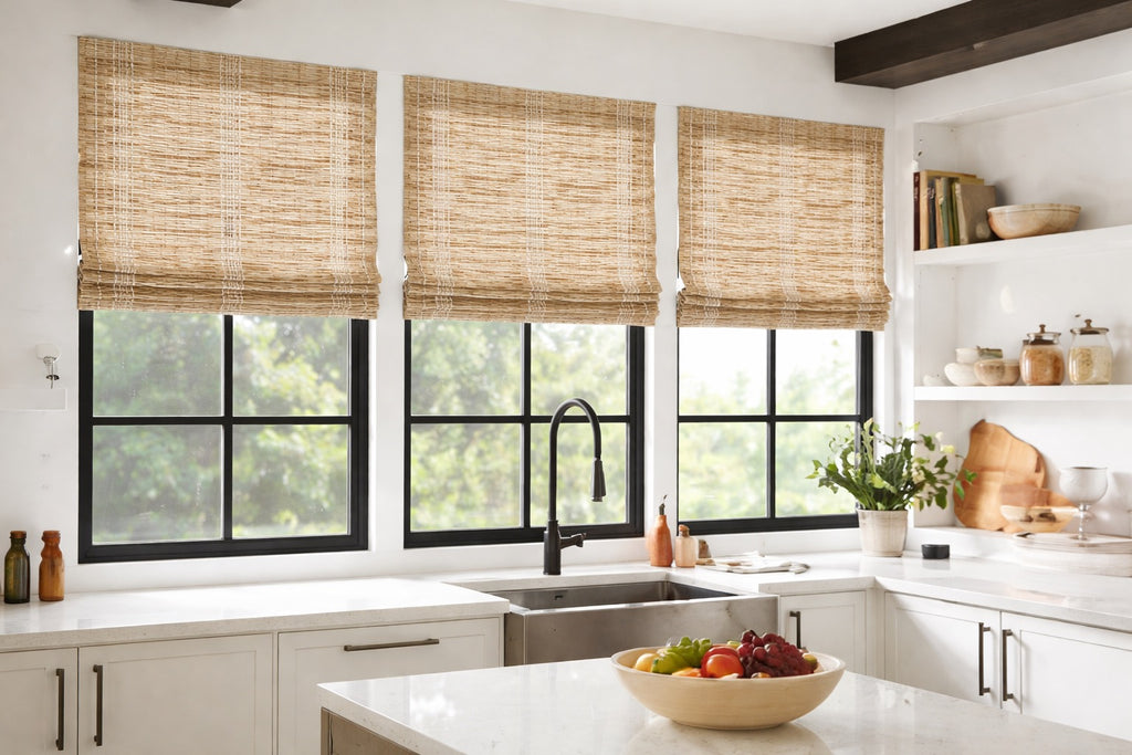 High-end kitchen with large black-framed windows featuring custom natural woven wood Roman shades in a warm neutral tone, styled above a farmhouse sink with white cabinetry, quartz countertops, and soft natural light.